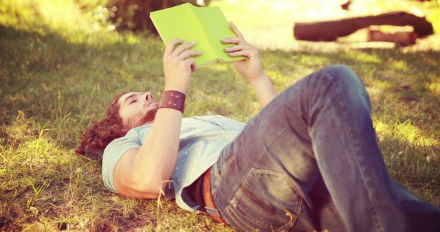 Young man lying on green grass while reading green book. Ideal for themes of leisure, relaxation, outdoor activities, literature or educational content. Can be used in advertisements for book clubs, libraries, outdoor recreation and wellness promotions.