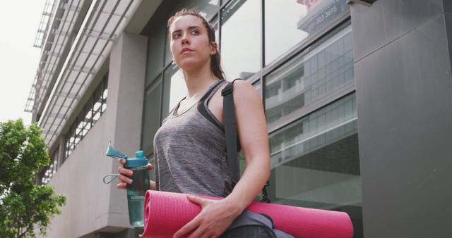 Young athletic woman wearing workout clothing holding pink yoga mat and water bottle, standing outside an urban office building with glass windows. She has a determined expression and looks ready for a workout or yoga session. Ideal for promoting fitness routines, yoga classes, urban workouts, and healthy lifestyle content.
