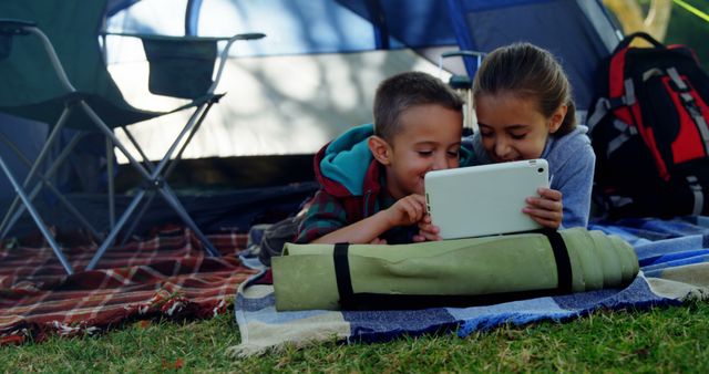 Two kids lay inside a tent, engrossed in a tablet, during a camping trip. This scene showcases the blend of technology and nature in modern outdoor adventures. Perfect for themes involving outdoor activities, family bonding, children's recreation, and tech-savvy vacations.