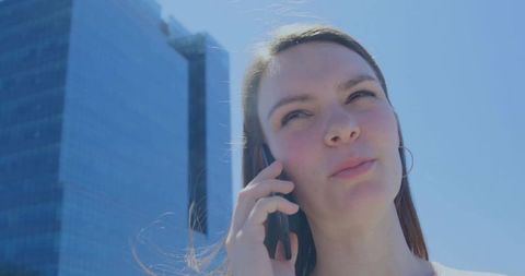 Young professional woman talking on smartphone near glass skyscrapers in sunlight