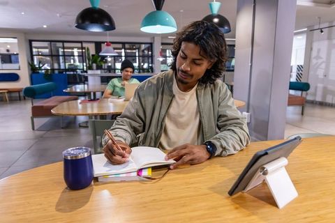 Indian man writing in notebook at coworking lounge with tablet and tumbler, modern workspace