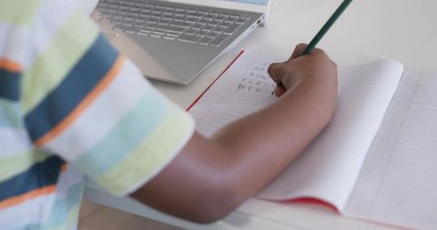 African american teen studying at desk writing math homework in notebook with laptop
