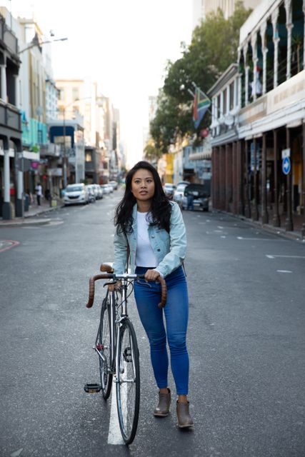 Young biracial woman walking with her bicycle on an urban street. She is wearing a denim jacket and jeans, looking at the camera. The street is relatively empty, suggesting early morning or a quiet time of day. This image can be used for themes related to urban lifestyle, transportation, city living, and casual fashion.