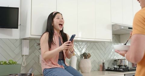 Surprised woman reacting to smartphone while partner stirring bowl in modern kitchen