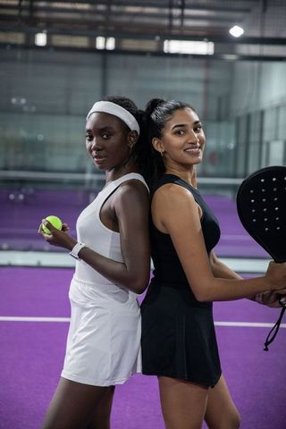 Diverse female padel partners standing back-to-back on purple court holding racket and ball