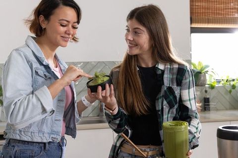Friends sharing homemade green smoothie bowl in bright kitchen, smiling and tasting