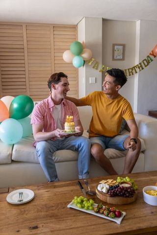 Two male friends celebrating birthday at home with cake, balloons, snacks and laughter