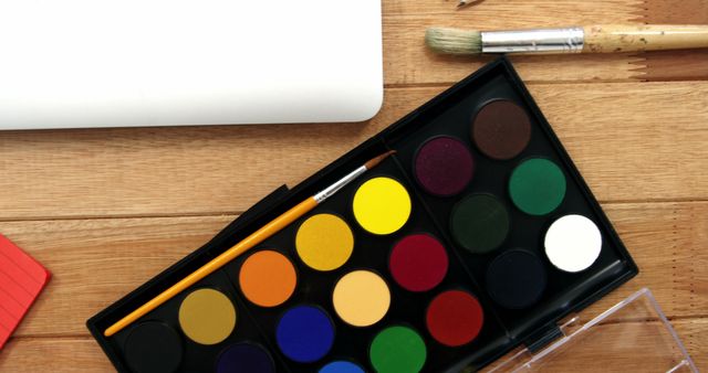 A watercolor paint set with a variety of colors lies next to brushes and a rolled-up piece of paper on a wooden surface, with copy space. It suggests a creative project or artistic endeavor about to begin.