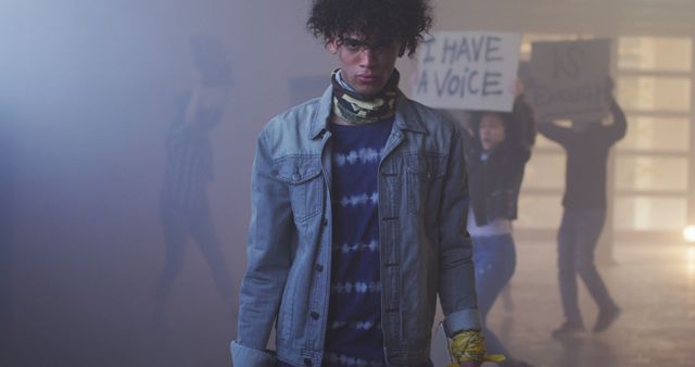 Young activist with curly hair in denim jacket stands firmly amid a smoky environment, flanked by others holding protest banners. The intense atmosphere highlights themes of social change, youth empowerment, and political engagement. Useful for articles, blogs, and campaigns on activism, youth movements, and social justice awareness.