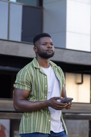 African american man standing by office facade holding smartphone looking thoughtful