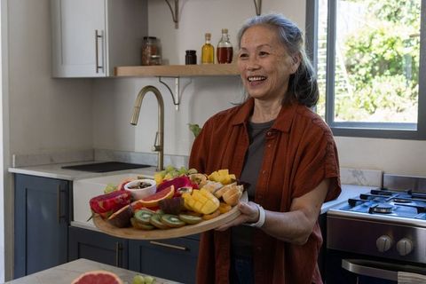Senior asian woman serving vibrant fruit platter in modern kitchen smiling hostess
