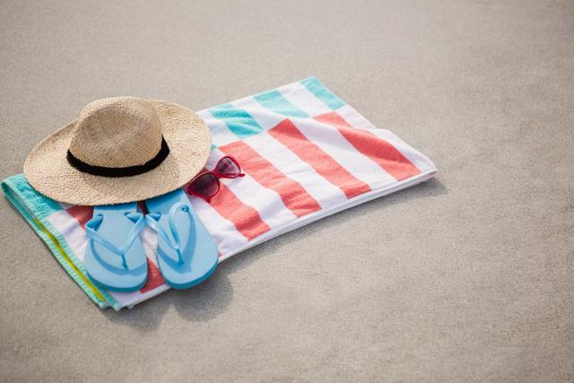 Perfect for travel and vacation-themed content, this image captures essential beach accessories including a straw hat, blue flip flops, and sunglasses placed on a colorful towel on sandy beach. Ideal for promoting summer holidays, beachwear, and relaxation.