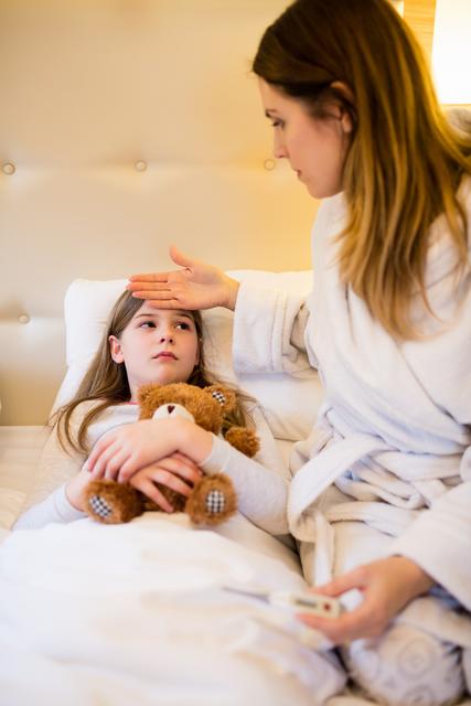 Mother checking her daughter's fever in a cozy bedroom. Daughter lying in bed holding a teddy bear, looking unwell. Mother showing concern and care. Ideal for use in articles about parenting, child health, family care, and home healthcare.