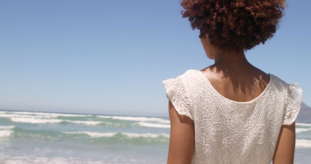 A woman with curly hair standing at the shoreline with a picturesque ocean background. Perfect for travel brochures, lifestyle blogs, wellness and relaxation promotions, summer vacation advertisements, and background designs for beach resorts.