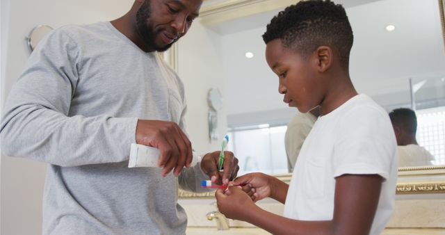 African american dad putting toothpaste on his son's brush in bathroom at home. fatherhood and love concept