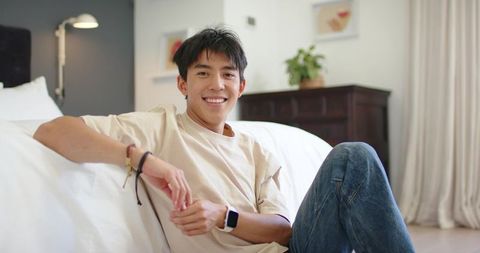 Smiling asian man relaxing on bed in minimal bedroom wearing smartwatch