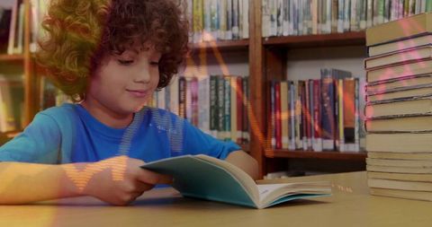 Curly-haired boy reading at library table wearing bright-blue shirt with stack of books