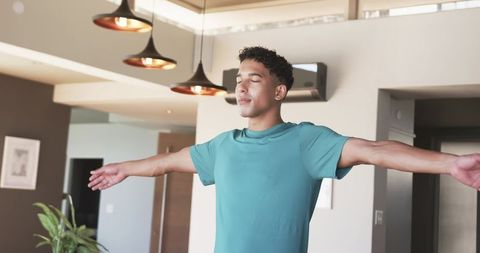Man standing with outstretched arms closing eyes wearing teal tee in modern apartment