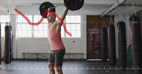 Man performing overhead barbell press in industrial gym with punching bags and analytics overlay