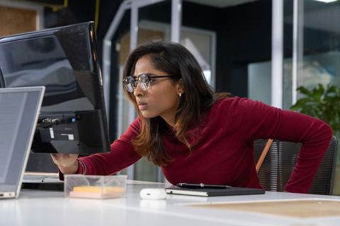 Indian young professional adjusting monitor while leaning at desk in open-plan office