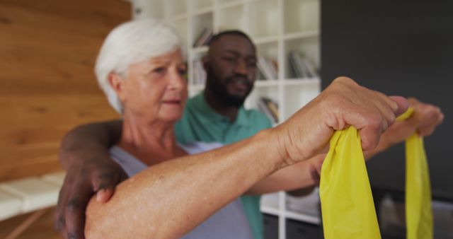 Senior woman using a yellow resistance band while guided by a trainer. Useful for illustrating physical therapy, elder care, fitness routines for seniors, or healthy aging. Appropriate for fitness blogs, healthcare websites, and senior wellness promotions.