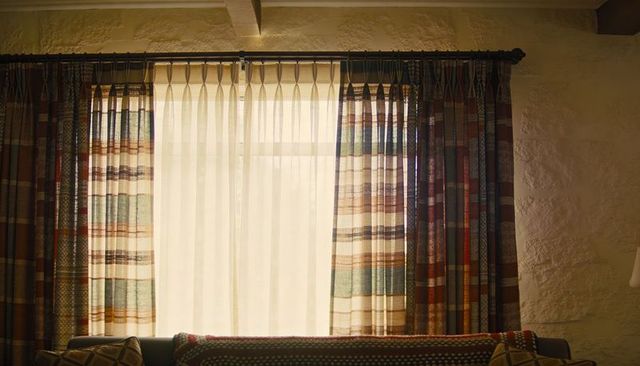 Framing sunlit living room window with sheer banded drapes, patterned curtains, rustic plaster wall