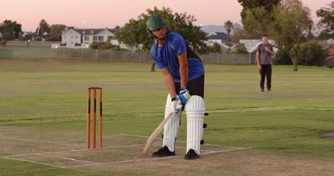 Young batsman practicing batting on cricket pitch at dusk wearing helmet and pads