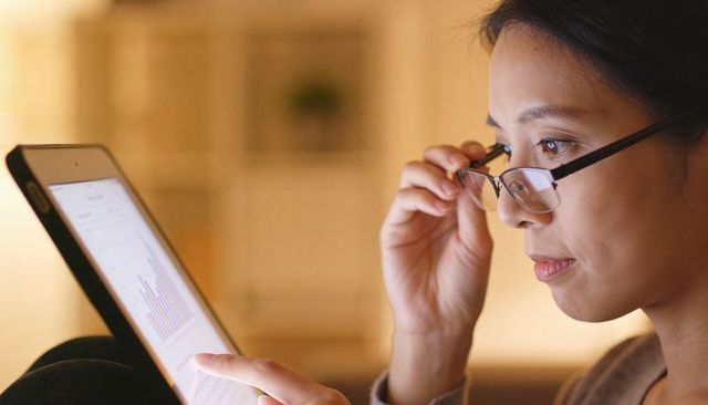 Woman analyzing bar chart on tablet while adjusting glasses in cozy living room evening