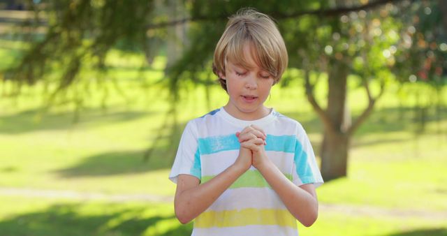 Young boy with hands clasped praying outdoors in a park surrounded by greenery. Useful for illustrating themes of childhood faith, spiritual moments, and the serenity of nature. Can be used in religious content, educational materials, and inspirational contexts.