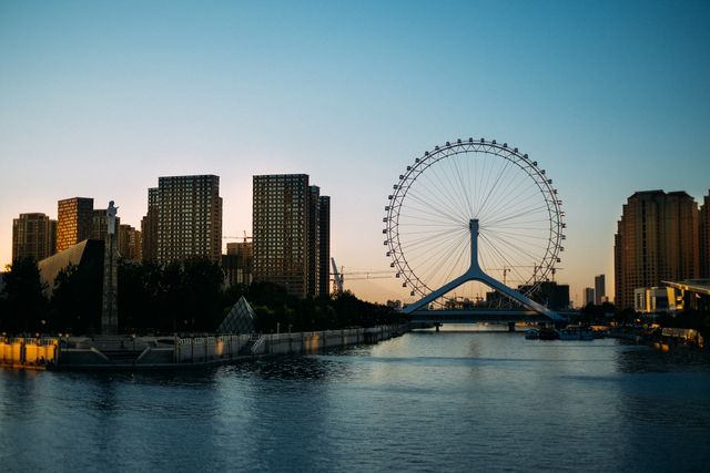 Skyline of a vibrant city features a prominent Ferris wheel and modern high-rise buildings against a sunset backdrop. Reflections on water amplify the urban beauty. Useful for depicting urban tourism, evening leisure activities, or city infrastructure.