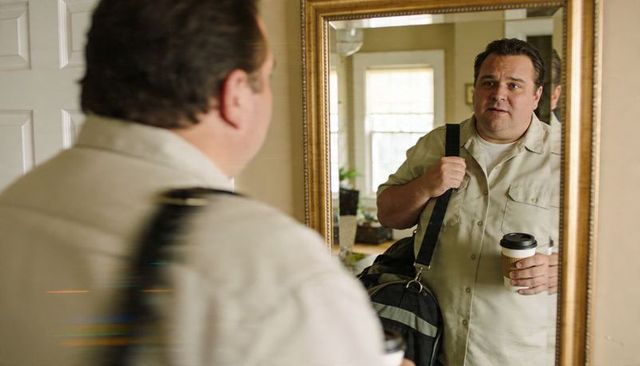 Middle-aged man checking mirror while holding duffel bag and takeaway coffee in foyer