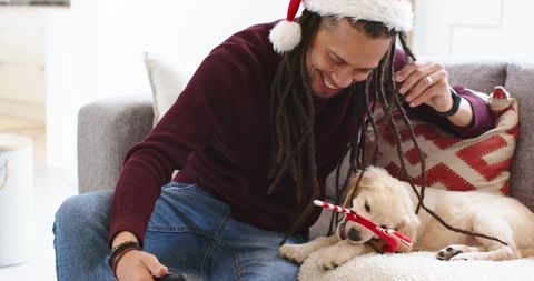 Mid-adult man wearing santa hat playing with puppy chewing candy cane on cozy sofa at home