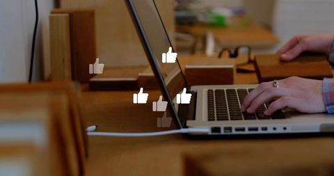 Hands typing on laptop with floating thumbs-up icons, wooden coworking desk, charging cable
