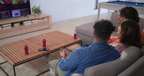 Group of friends relaxing on sectional sofa watching streaming tv at home with bottled drinks