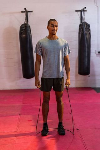 African american man jumping rope in boxing gym on red mats with heavy bags for cardio