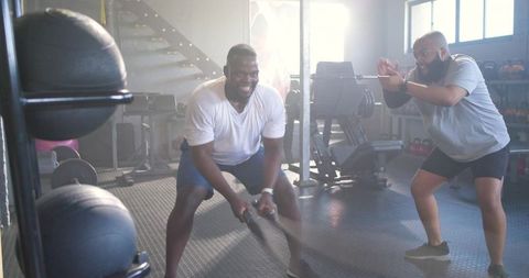 African american men using battle ropes, trainer coaching high-energy gym workout
