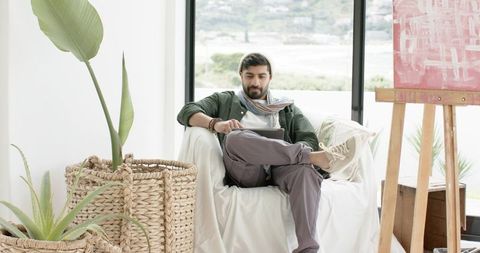 Indian man relaxing in bright studio using tablet near waterfront window, canvas, plants