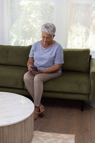Senior woman sitting on olive green sofa using smartphone for messaging and browsing
