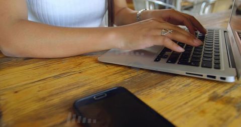 Asian woman typing on silver laptop at wooden cafe table with smartphone, remote work