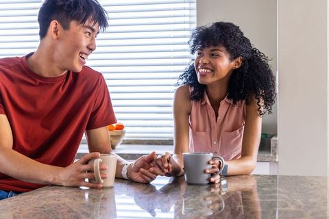 Young couple sharing coffee and laughing while holding hands at kitchen countertop