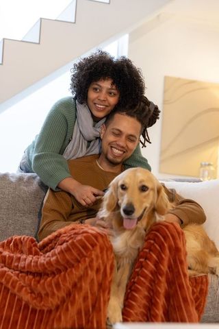 African american couple cuddling and smiling on sofa with golden retriever and blanket