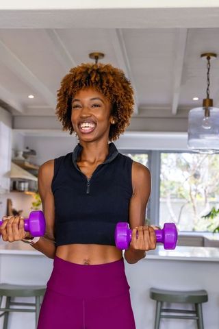 African american woman lifting purple dumbbells in kitchen smiling during home workout