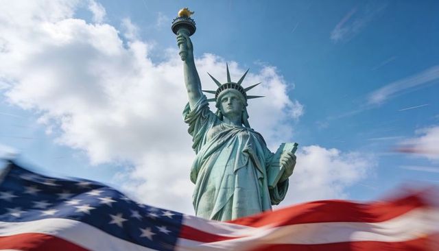 Statue of liberty holding torch and tablet with waving american flag against blue sky