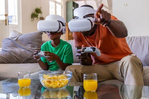 African American father and son playing VR together on living room sofa with controllers and snacks