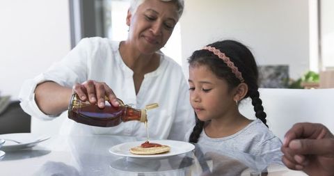 Happy grandmother serving pancakes to young granddaughter with syrup