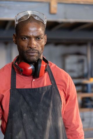 African american craftsman standing in workshop wearing safety goggles, ear protectors, apron