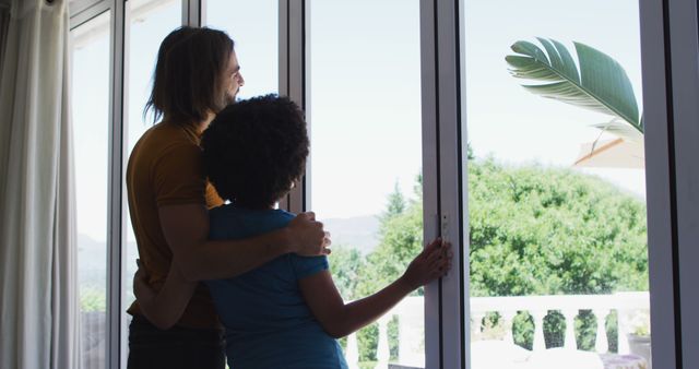 Couple standing close together and looking out large windows at a view of greenery and clear sky. Ideal for home living, relationships, diversity, and lifestyle themes. Can be used for real estate, wellness, and family-oriented advertisements.