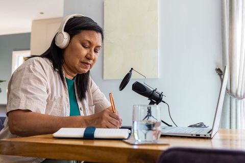 Asian woman recording podcast at home office wearing headphones and taking notes with laptop and mic