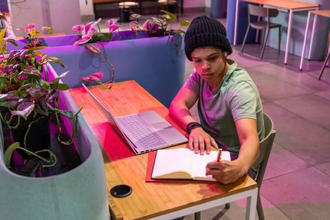 Young man writing in notebook at modern coworking cafe with laptop and neon lighting