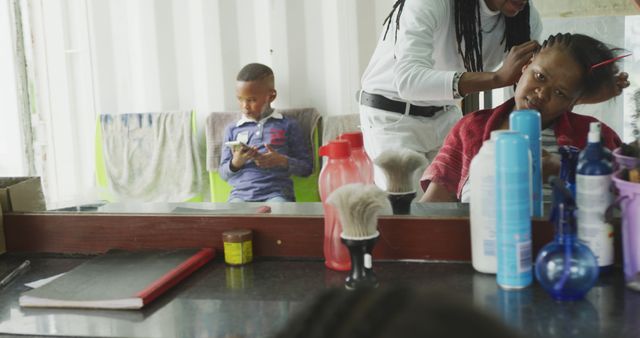 Young boy reading book in a barbershop while stylist braids another child's hair. Suitable for illustrating themes of hair care, grooming, education, multitasking, or childhood activities. Can be used in advertisements or websites related to barbershops, hair salons, educational content, family life, or cultural representations.